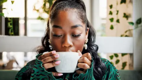 Front view of a woman with pink nails and green jacket sipping from a white cup.