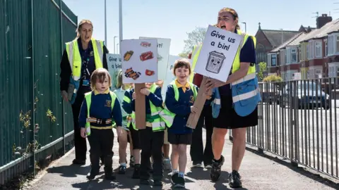 Young boys and girls in their blue and yellow school uniform hold up placards saying "Give us a bin!" as they march on a pavement, between railings on both sides. They are accompanied by two female school staff with hi-vis jackets.