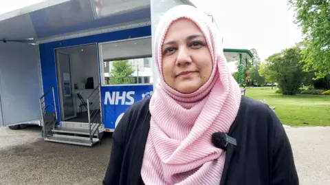 Mark Norman BBC Dr Samia Kidwai looks at the camera with an NHS mobile vaccination centre in the background.