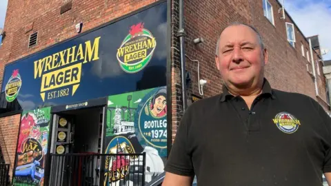 Mark Roberts of Wrexham Lager, wearing a company-branded shirt, stands outside the business, which is also branded with company logos and posters