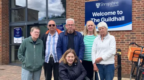6 people stand outside a brick building with blue and white signage saying 'Shropshire Council' 'Welcome to the Guildhall' . A person in a green jacket and glasses stands on the outside, next to a man in sunglasses and a striped blue and white shirt. Next to him is a man with white hair and glasses in a blue coat, holding on to the wheelchair of the lady at the bottom of the picture who has brown hair and a navy jacket on. Next to them are two women, one is blonde with a green and white striped top and a lady with glasses and a white top. They all look unhappy. 