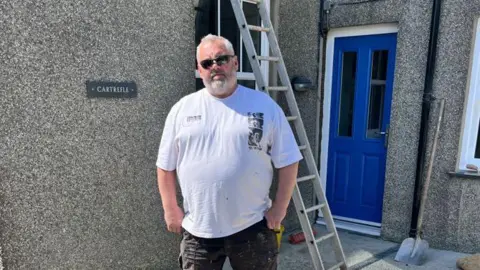 Jason is wearing a white t-shirt and black shorts. He has grey hair and beard and is wearing black sunglasses. He is stood in front of a pebble-dashed house. It has a blue door and a ladder and shovel in view behind him.