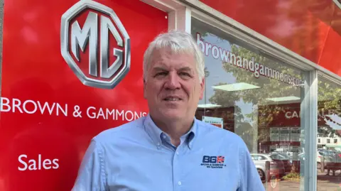 Tony Fisher/BBC A man with grey hair and wearing a blue shirt with a logo on it stands outside an MG car showroom which is called Brown & Gammons. The large window behind him reflects some cars parked outside.