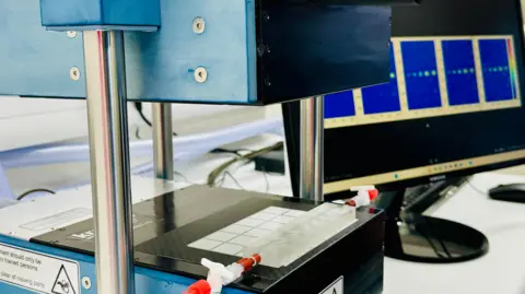 The prototype machine is blue and has a top and bottom part which will compress the breast. Next to it is a tray where radioactive tracers have been placed.
On the computer screen beside the machine you can see a row of small lights - which demonstrate how the machine can 'light up' cancerous tumours in the breast.