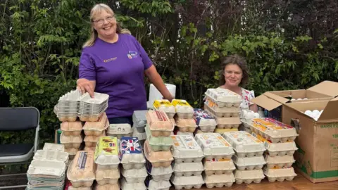 Towcester Community Larder Two women are behind a table with a large collection of egg boxes on it. The woman on the right is sitting down. The woman on the left is standing up and wearing a purple T-shirt. A chair is to the left of the table and a cardboard box with white eggs in it, to the right. There are trees in the background. 