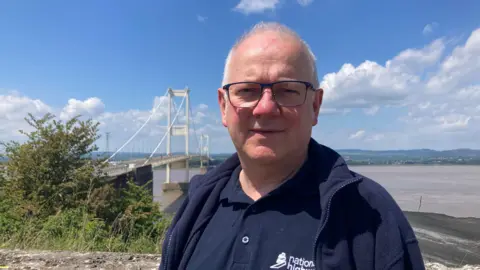 Chris, wearing glasses and a navy blue polo shirt and fleece, stands with the M48 Severn Bridge in the background. It is a clear day with blue sky and some cloud.