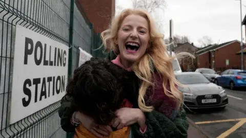 PA Media Hannah Spencer stands in front of a polling station sign attached to a metal fence. Spencer is smiling, holding an orange coloured bag and wearing a multi-coloured coat.