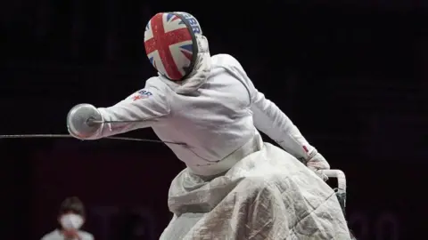 Dimitri Coutya leaning out of his wheelchair towards his opponent to strike their torso with his fencing foil (sword). He is dressed in all white protective kit with gloves, a lap cover and a fencing hood, which has the Great British flag on it. There is also the Great Britain logo on his shoulder.