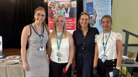 Supplied Hannah Popple (PCN Manager), Sarah Chappell (Redmoor Health Programme Manager), Dr Karen Forshaw (GP and Clinical Director), Zara Bibi (Pharmacy Technician) stood together with their lanyards in front of their health care banners.