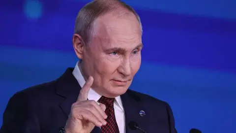 Russian President Vladimir Putin from chest up, holding one finger up as he delivers a speech 