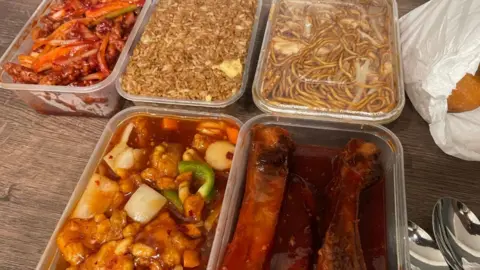 Plastic takeaway containers filled with Chinese food arranged on a wooden table: fried rice, noodles in sauce, sweet-and-sour chicken with vegetables, ribs in dark sauce, and a spicy stir-fry, with a white and metal spoons nearby.