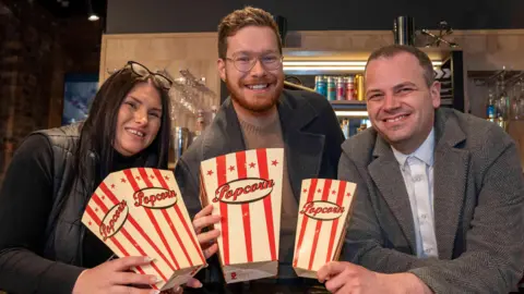 Lisa Roberts, Lockworks Cinema assistant manager, councillor Chris Burden and Darryl Griffiths, Lockworks cinema manager lean over a bar at the cinema, holding red and white popcorn boxes. 