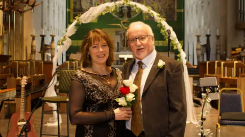 A woman and man stood in front of a white, floral arch at Coventry Cathedral. The woman is wearing a black outifit with silver embroidery and holding red and white flowers. The man, who has glasses and grey hair, has a brown suit and tie with white shirt