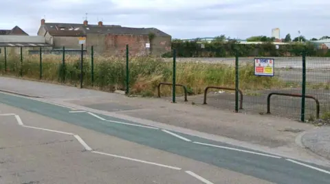 Empty land behind fence with hard-standing and weeds growing. A road and pavement in the foreground and red-brick buildings in the background.