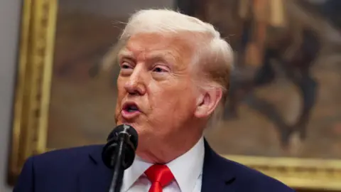 Donald Trump wearing a blue suit, white shirt and red tie, with a gold-framed painting in the background, speaking at the White House 