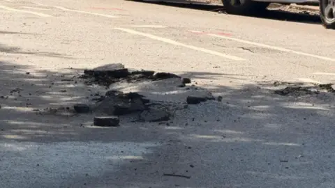 The image shows a section of road that was hit by lightning, it has damaged the tarmac and left it very uneven with rubble around it.