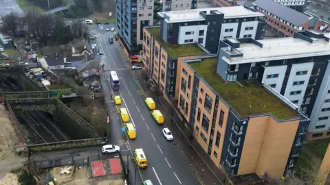 BBC/Oli Constable An aerial view of an urban area featuring a wide road running through the centre of the scene. Along this road, several yellow emergency vehicles, likely ambulances, are lined up in a row, parked on one side of the street. A red emergency vehicle, possibly a fire engine, is also visible further up the road near an intersection. A white bus is stopped in the middle of the road, and a few other cars are nearby.