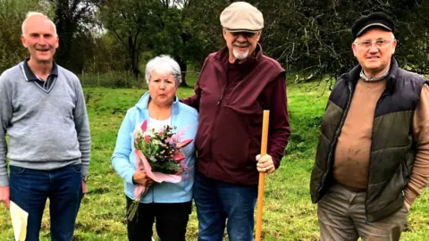 Barton Family Four older people standing in a field and looking at the camera. The man on the far left is wearing dark blue jeans and a grey v-neck jumper with a black polo shirt underneath. He has white hair and is balding. In the middle is a man who has his arm around a woman. He is wearing a maroon jacket and beige flat cap and has a white beard. The woman has short grey hair, is wearing a light blue zip up top and is holding a bunch of flowers. The man on the right is wearing beige trousers, with a brown gilet and an orange/brown jumper.