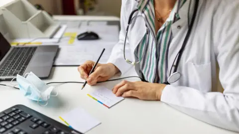 A generic image of a caucasian female GP in a white lab coat and striped purple and green shirt writing at a desk. She has a stethescope around her neck. 