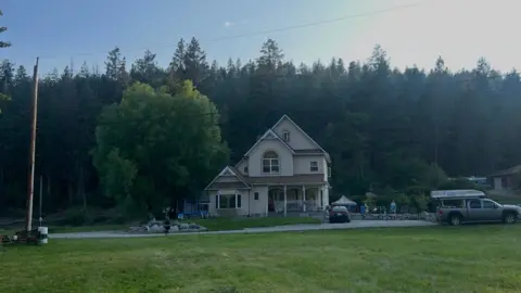 Supplied/Keegan Radomski An image showing Keegan Radomski's home - a two-storey house with a slanted roof and an attic, a drive way with two cars visible parked nearby. Behind it is a row of large spruce trees. In front of it is a large grass field. 