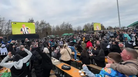 The Newcastle United fan zone - hundreds of people standing, with many raising their arms into the air in response to a goal. Two large screens depict player Dan Burns running down the pitch after scoring