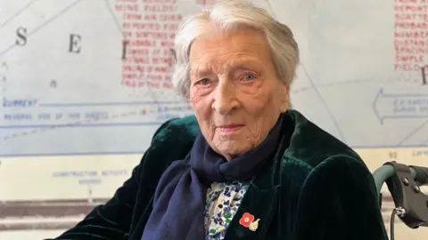 Christian wearing a velvet jacket and a scarf with white hair and blue eyes smiling at the camera. She is in a wheelchair and you can see a map in the background but it is not in focus.