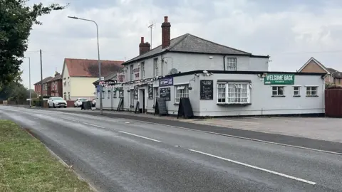 Richard Knights/BBC The road outside the Cherry Tree pub, which is a grey and black building with a small car park to its side.