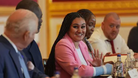 PA Media Serena Wiebe is wearing a pink blazer with a white T-shirt underneath. She is sitting at a long table wearing a blue cast on her right arm. Serena is smiling at King Charles, who is facing her, not the camera. Idris Elba sits in between the King and Serena. 