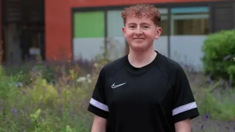 Sheffield Children's Hospital Harrison Sherwell, with short brown curly hair is wearing a black casual fit t-shirt with a thick white horizontal stripe on the sleeves. He is smiling, standing against a backdrop of wildflowers.