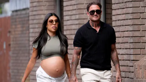 PA Media Stephen Bear (right) holding hands with Miami (left) as they walk into court, with a brick wall behind them. Miami is heavily pregnant with her bump showing and she has long black hair and big sunglasses on, and white trousers.