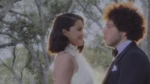 Selena Gomez via Instagram Selena Gomez, a young woman with dark hair, wears a sleeveless, high-necked white wedding gown and is embracing Benny Blanco, a young man wearing a dark tuxedo and white shirt. They are standing in front of trees.