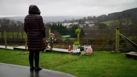 A woman in a burgundy jacket, with her hood up, looking at a grave on the edge of a cemetery. In the background is a view of trees, fields and houses.