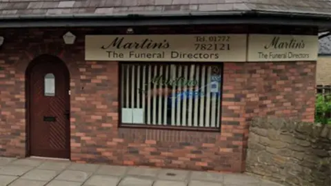 Streetview image of the funeral directors in Longridge. A red brick building with a brown arched door to the left and a window to the right with a vertical cream blind.