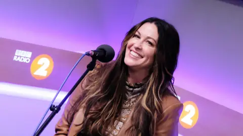 BBC A smiling lady with long dark hair and a brown leather jacket, talking into a microphone