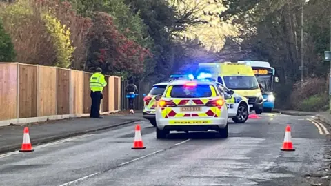 Two police cars and an ambulance blocking a road. There are three small orange and white traffic cones blocking the road in the foreground. A police officer in yellow high-vis coat is stood on the path on the left of the image. A bus can be seen facing the camera in the background behind the emergency vehicles.