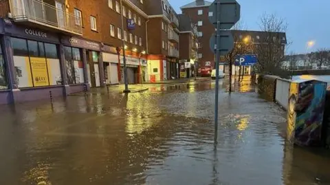 RTE Flooding in Cork City has turned a city street into a shallow swamp of perhaps half a foot. The water is high enough to cover the very bottom of signposts, shop fronts and what appears to be the outer wall that lines a river. 