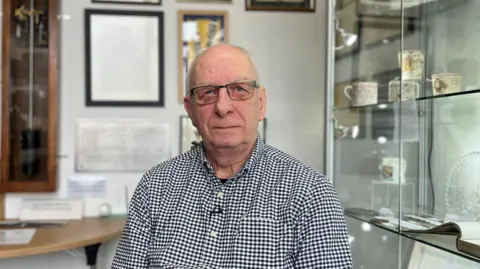 Paul Roper is wearing a blue and white check long sleeved shirt and glasses and is looking at the camera inside the Torpoint Archive room at Torpoint library. Beside him are display cabinets containing documents, mugs and glassware. On the wall behind are framed documents and the bottom part of a clock in a case