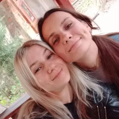 Supplied The photo shows two women smiling at the camera. The woman on the left has long blonde hair, and the woman on the right has brown hair. Their heads are pressed together, and they are sitting in front of what appears to be a green garden.