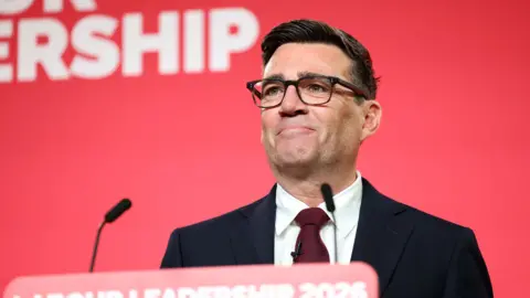 Andy Burnham smiling  and standing in front of a red backdrop  as  the newly announced leader of the Labour Party makes, his first speech to party members in central London on July 17, 2026 in London, England.
