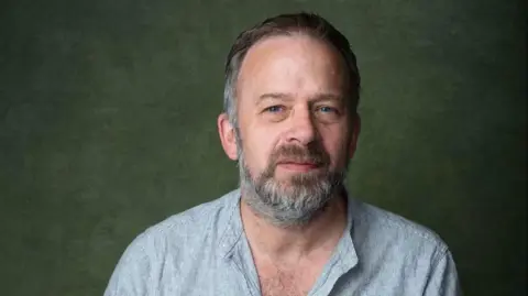 The Pound Arts Centre Russ Tunney is looking into the camera with a slight smile. His hair and beard is greying. He is wearing a shirt with a fine blue and white stripe.