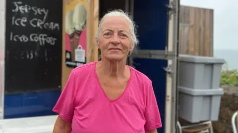 Mary is wearing a bright pink T-shirt stands in front of a small wooden refreshment kiosk near the coast. A blackboard sign on the kiosk advertises “Jersey ice cream” and “Iced coffee.” Storage containers, folded chairs, and a glimpse of the sea are visible in the background.