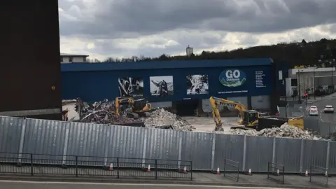 Local Democracy Reporting Service Two diggers tearing down a former retail site surrounded by rubble. The remaining building is blue with a Go Outdoors logo on the side.