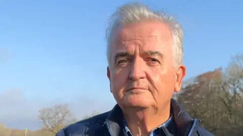 Councillor Leslie Rice is outside near the golf course. The close-up photograph shows him standing under a clear blue sky with trees behind him. He has grey hair and is wearing a navy jacket.