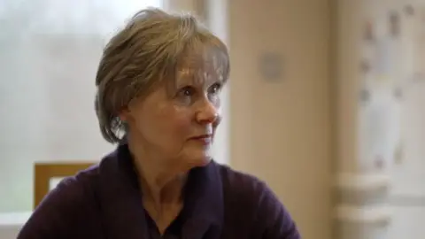 Andrea looks to the side as she is interviewed by the BBC. She is wearing a navy blue blouse and has short light brown hair. A window to the countryside can be seen in the background.