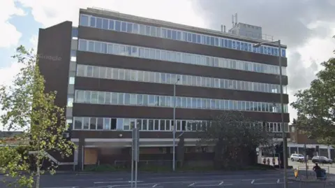 A large office block, six storeys high with lots of windows and bleak brown walls.