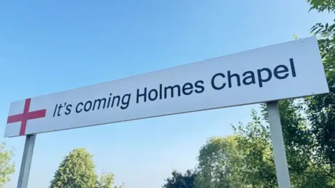 Northern Railway station sign reading: 'It's coming Holmes Chapel'.