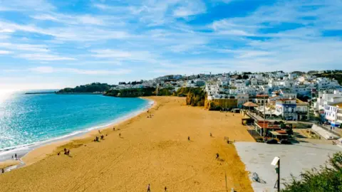 A view of the Albufeira coastline in Portugal. People enjoy the sand beach which is overlooked by white buildings.
