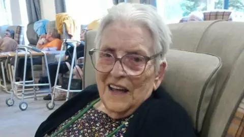Family handout Lisa Greenwood's mother Mavis is pictured smiling at the camera in a care home setting. She is wearing glasses, and a dark cardigan over a dress. 