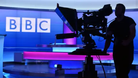 A cameraman films a blue and pink studio showing the BBC logo 