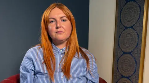 Kirsty Haynes sits on a red velvet sofa with a mandala decoration on the wall. Kirsty has red long hair and blue eyes. She is wearing a blue and white striped shirt. 